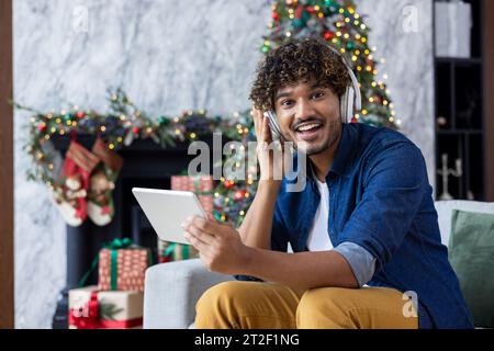Porträt eines jungen hispanischen Mannes, der während der Winterferien zu Hause auf dem Sofa neben dem Weihnachtsbaum sitzt. Mit Kopfhörern hört er Musik von einem Tablet, ruht sich aus, lächelt in die Kamera. Stockfoto
