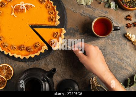 Zugeschnittene Ansicht eines Mannes, der ein Stück Danksgiving Kürbiskuchen in der Nähe einer schwarzen Teekanne auf einem rauen Steintisch nimmt Stockfoto