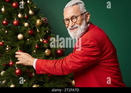 Fröhlicher Mann, gekleidet als stilvoller Weihnachtsmann in Gläsern, die roten Marmor auf Tanne berühren, Winterkonzept Stockfoto