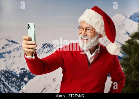 Glücklicher Mann, der als Weihnachtsmann gekleidet ist, Brille trägt und Selfie mit Bergkulisse macht, Winterkonzept Stockfoto