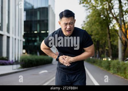 Asiatischer junger männlicher Sportler beugte sich vor Schmerzen in der Seite und hielt seinen Bauch mit den Händen, nachdem er durch die Stadt gelaufen war. Stockfoto