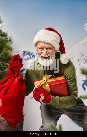 Fröhlicher Weihnachtsmann mit weißem Bart in weihnachtlicher Mütze mit Geschenktasche und Geschenk in der Hand, Winterkonzept Stockfoto