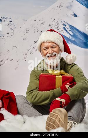 Fröhlicher, weißbärtiger Mann, gekleidet als Weihnachtsmann, sitzend auf Schnee mit Geschenk in den Händen, Winterkonzept Stockfoto