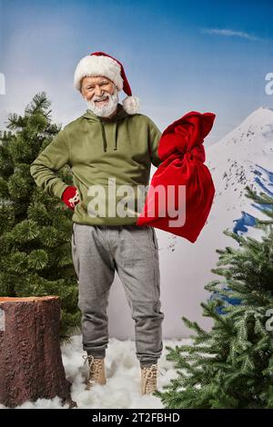 Fröhlicher Mann in khakifarbenem Kapuzenpulli, der die Weihnachtsmanntasche hält und weihnachtliche Mütze trägt. Frohe Weihnachten Stockfoto