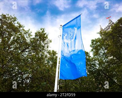 Das Logo der Internationalen Atomenergie-Organisation (IAEO) auf einer blauen Flagge neben Bäumen vor dem Queen Elizabeth II Centre in London, Vereinigtes Königreich. Stockfoto