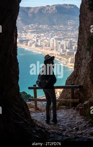 Ein junger Wanderer mit Hut im Tunnel des Naturparks Penon de Ifach mit der Stadt Calpe im Hintergrund, Valencia. Spanien. Mittelmeer Stockfoto