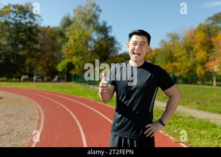Porträt eines lächelnden jungen asiatischen Mannes, der in Sportbekleidung in einem Stadion steht und Superfinger auf die Kamera zeigt. Stockfoto
