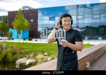 Glücklicher junger asiatischer Mann, der Sport treibt und in der Nähe des Sees in der Stadt läuft, Kopfhörer trägt und ein Telefon hält. Stockfoto