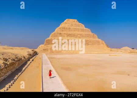 Eine junge Frau in rotem Kleid besucht die Stufenpyramide von Djoser, Sakkara. Ägypten. Die wichtigste Nekropole in Memphis. Die erste Pyramide in Stockfoto