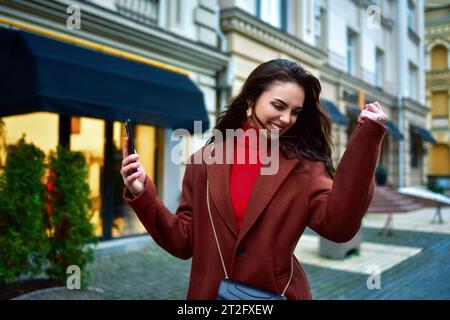 Schöne überfröhliche junge Brünette Frau mit langen Haaren in einem roten Mantel, die gute Nachrichten feiert, Telefon hält und Faust in der Abendstraße geballt Stockfoto