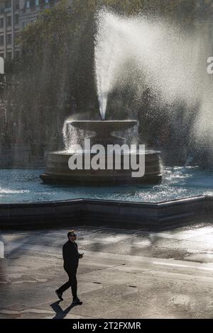 Mann, der an einem der Springbrunnen am Trafalgar Square, London, an einem windigen Herbsttag vorbeiläuft Stockfoto
