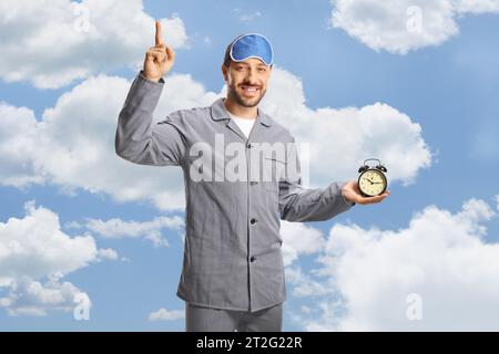 Mann im Pyjama mit Schlafmaske, der einen Wecker hält und auf die Wolken zeigt Stockfoto