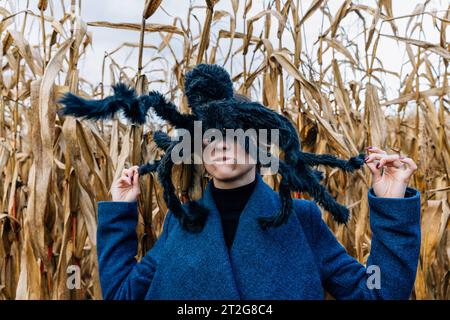Riesige schwarze Spielzeugspinne auf dem Kopf einer Frau auf einem Maisfeld während einer Halloween-Party auf einem Maisfeld Stockfoto