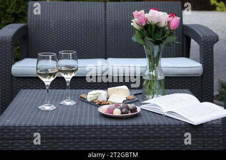 Vase mit Rosen, Gläsern Wein, offenem Buch und verschiedenen Snacks auf Rattantisch auf der Außenterrasse Stockfoto