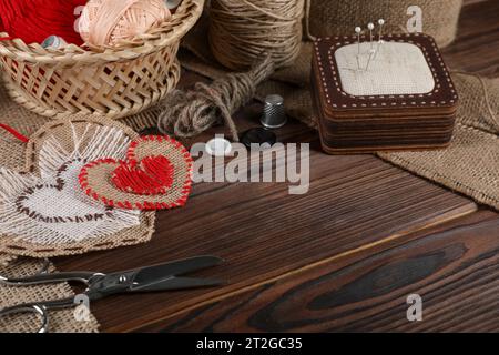 Herzförmige Stücke aus Sackleinen-Stoff mit verschiedenen Stichen und Nähwerkzeugen auf Holztisch, Nahaufnahme. Leerzeichen für Text Stockfoto