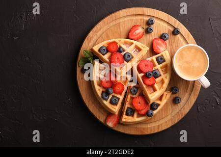 Leckere belgische Waffeln mit frischen Beeren und einer Tasse Kaffee auf schwarzem Tisch, Blick von oben. Leerzeichen für Text Stockfoto