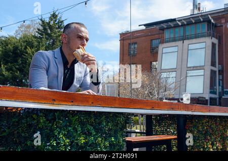 Kaukasischer Geschäftsmann mit Jacke, Sandwich zum Frühstück, Essen, Ausblick und Nachdenken. An der Bar vor dem Restaurant sitzen, Stockfoto