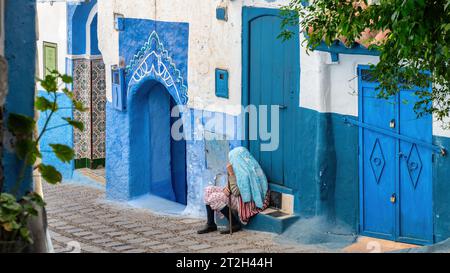 Chefchaouen, Marokko - 11. September 2022: Alte Frau sitzt bei ihrem Haus in Chefchaouen, bekannt als die blaue Stadt, berühmt für ihre auffällige blaue Farbe PA Stockfoto