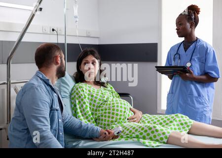 Zukünftige Eltern diskutieren mit Assistentin in der Krankenhausstation, schwangere Frau, die im Bett liegt und Wehen hat. afroamerikanische Krankenschwester, die Patientin mit Schwangerschaft auf die Operation vorbereitet Stockfoto