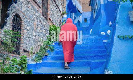 Chefchaouen, Marokko - 11. September 2022: Frau in einem roten burnu, die auf den blauen Straßen von Chefchaouen läuft, bekannt als die blaue Stadt, berühmt für ihre s Stockfoto