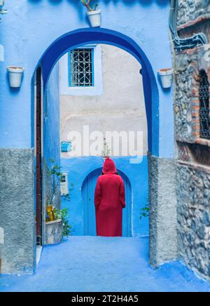 Chefchaouen, Marokko - 11. September 2022: Frau in einem roten burnu, die auf den blauen Straßen von Chefchaouen läuft, bekannt als die blaue Stadt, berühmt für ihre s Stockfoto