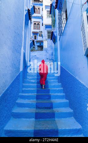 Chefchaouen, Marokko - 11. September 2022: Frau in einem roten burnu, die auf den blauen Straßen von Chefchaouen läuft, bekannt als die blaue Stadt, berühmt für ihre s Stockfoto