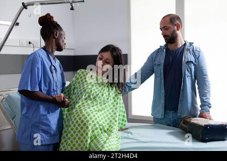 afroamerikanische Assistentin hilft schwangeren Frauen, aus dem Bett in der Krankenhausstation zu kommen und bereitet den Patienten auf eine Kaiserschnittoperation vor. Zukünftige Eltern, die ein Kind in der Entbindungsklinik erwarten Stockfoto