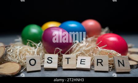 Farbenfrohe Ostereier liegen im Strohhalm, umgeben von Holzdekorationen. Ostervorbereitungen. Auf dem Tisch stehen Buchstaben, die das Wort ostern beschreiben Stockfoto