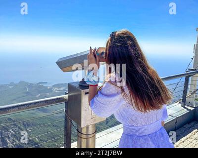 Ein Mädchen in einem weißen Kleid mit langen Haaren schaut durch ein Fernglas auf einer Aussichtsplattform. Besichtigungstour, neugieriger Tourist. Stockfoto