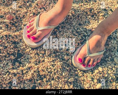 Ein Mädchen mit einer pinkfarbenen Pediküre an den Füßen spaziert in einem warmen Land in weißen Gummischuhen. Freizeitschuhe stehen auf dem Sand. Schieferplatten zum Gehen entlang der Stockfoto