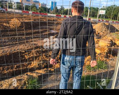 Ein junger starker Kerl steht hinter einem Metallzaun und hält ihn fest. Der Typ kletterte über die Bars, um zur Baustelle zu kommen, jetzt versteckt er sich vor Stockfoto