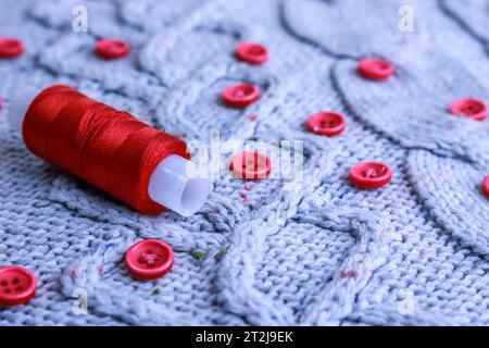 Schöne Textur eines weichen, warmen Naturpullovers, Stoffe mit einem Strickmuster aus Garn und roten kleinen runden Knöpfen zum Nähen und einem Knäuel aus rotem Thr Stockfoto