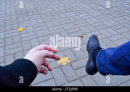 Ein Mann hält eine Zigarette in den Händen, Finger sitzen im Park und rauchen mit seinem Bein in einem Schuh vor dem Hintergrund von Pflasterplatten mit Autum Stockfoto