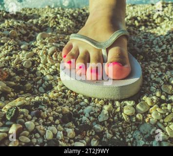 Ein Mädchen mit großen Fingern und einer pinkfarbenen Pediküre an den Füßen spaziert in einem warmen Land in weißen Gummischuhen. Freizeitschuhe stehen auf dem Sand. Schiefer f Stockfoto