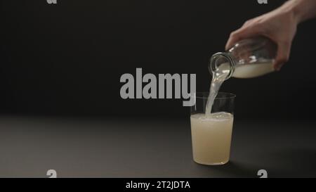 Man Hand gießen Zitrone Ingwer Limonade in Glas auf schwarzem Papier Hintergrund mit Kopierraum, breites Foto Stockfoto