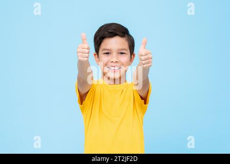 Glücklicher gemischter Race Boy in gelbem T-Shirt lächelnd und in die Kamera schauend, während er die Daumen nach oben zeigt, die Geste der Genehmigung vor blauem Hintergrund Stockfoto