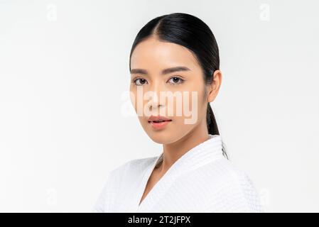 Hübsche junge asiatische Frau im Bademantel mit sauberer Gesichtshaut, die während des Spa-Kurses vor weißem Hintergrund in die Kamera schaut Stockfoto
