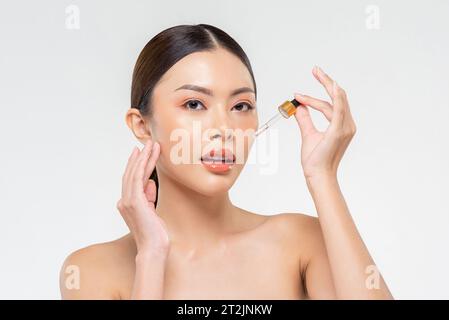 Beauty-shot-Porträt einer attraktiven asiatischen Frau, die Hautpflegeprodukt auf Gesicht von Pipette in weißem isoliertem Hintergrund aufträgt Stockfoto