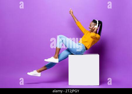 Lächelnde junge afroamerikanische Frau mit Kopfhörern, die auf dem Hocker sitzen und Musik hören und den Arm heben, im violetten Studio-Hintergrund Stockfoto