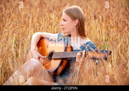 Schöne junge Frau, die im Weizenfeld sitzt und Akustikgitarre spielt. Stockfoto