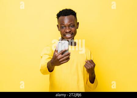 Aufgeregt junger afrikanischer Mann, der die Faust geballt hat und auf das Handy auf isoliertem gelbem Studiohintergrund blickt Stockfoto