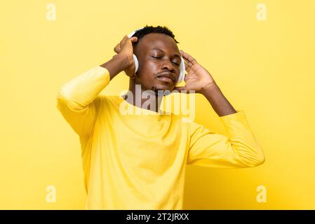 Junger afrikanischer Mann, der mit geschlossenen Augen Musik hört, während er Kopfhörer vor gelbem Studiohintergrund hält Stockfoto