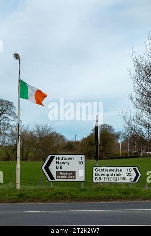 Die Flagge der Republik Irland fliegt über Straßenschildern, mit skurrilen Graffitis, die für einen lokalen Pub außerhalb von Hilltown, Co. Werben. Nach Unten Stockfoto