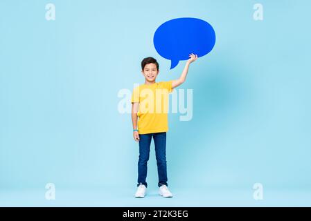 Asiatischer Junge mit gemischter Rasse, der legere Kleidung trägt, steht mit einer Hand haltenden Sprechblase auf hellblauem, isoliertem schwarzem Atelier Stockfoto