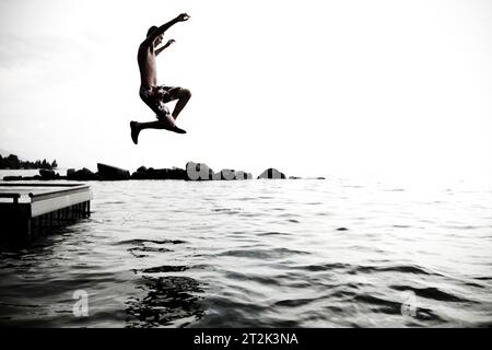 Ein junger Teenager wirft seine Arme in die Luft, während er von einem Dock springt, um im kühlen Wasser des Flathead Lake, Montana, zu schwimmen. Stockfoto