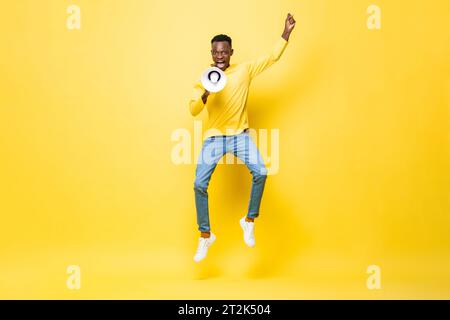 Mächtiger energischer junger afrikanischer Mann, der auf Megaphon in Studiogelb springt und brüllt Stockfoto