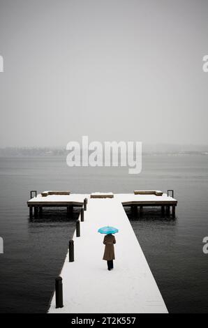 Eine junge Frau läuft auf einem schneebedeckten Dock mit Blick auf einen See, während sie einen hellblauen Regenschirm hält. Stockfoto