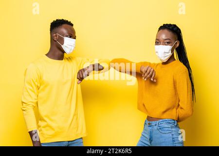 Das junge afroamerikanische Paar trägt Gesichtsmasken und stößt sich während der COVID-19-Pandemie in einem isolierten gelben Studiohintergrund an die Ellbogen, um sie zu begrüßen Stockfoto