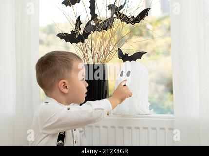 Halloween-Konzept. Keine Fläche. Die Hände des Babys in der Nähe des Fensters, vor dem Hintergrund einer Vase mit trockenen Zweigen und schwarzen Papierfledermäusen. Nahaufnahme Stockfoto