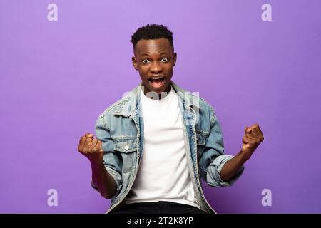Er gewinnt den ekstatischen jungen afrikanischen Mann, der seine Fäuste mit aufgeregtem Gesicht in isoliertem lila Farb-Studio-Hintergrund geballt Stockfoto